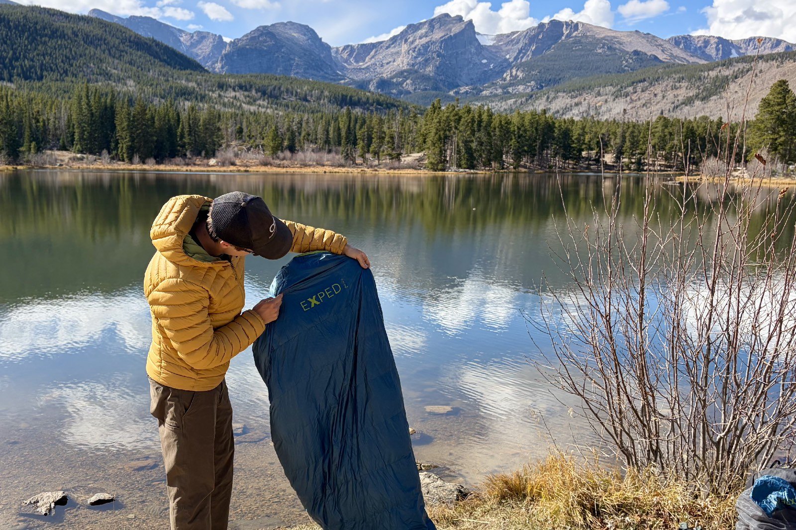 a hiker holds a blue sleeping bag next to a mountain lake