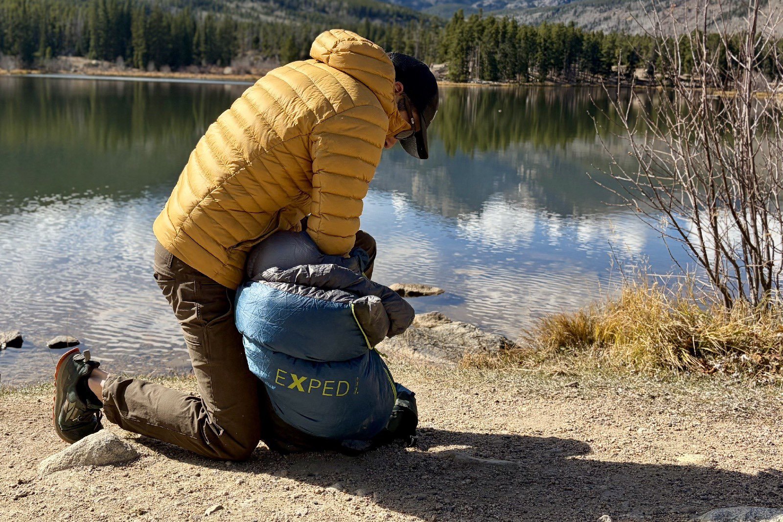 a hiker stuffs a blue sleeping bag into its sack on the shore of a lake
