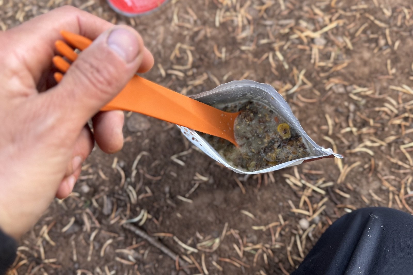 A POV view of a hiker dipping a plastic spork into a backpacking meal on the ground