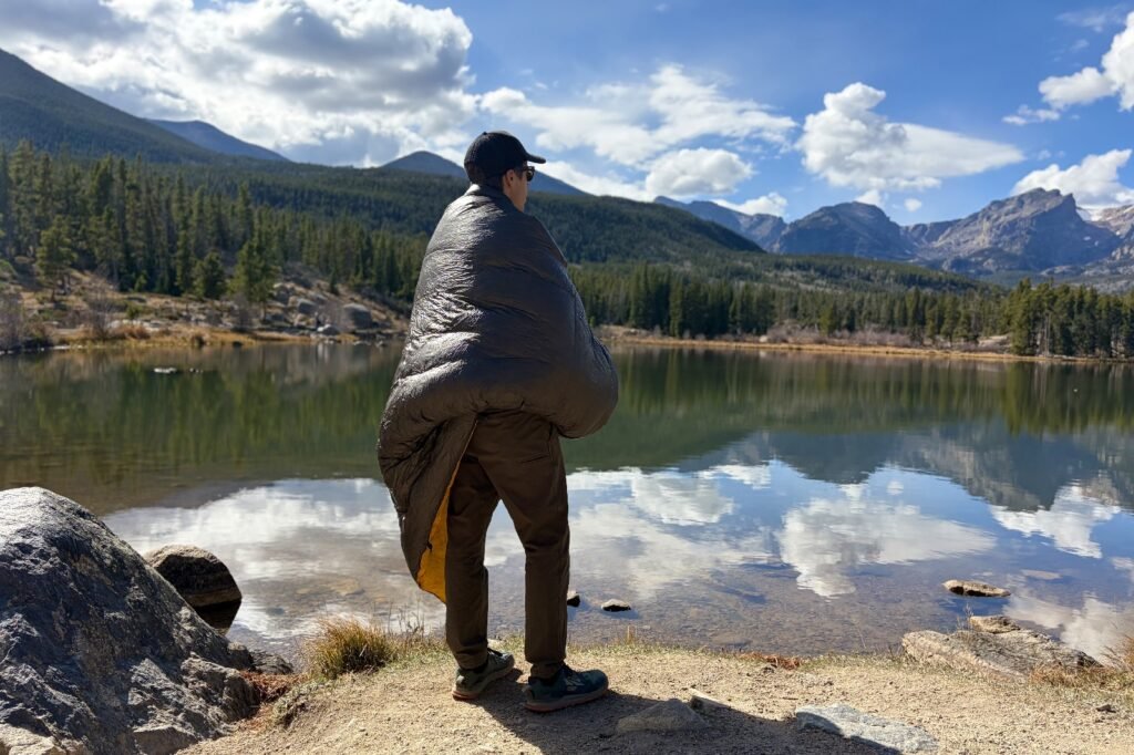 a hiker stands wrapped in a grey quilt looking over a lake and mountain view
