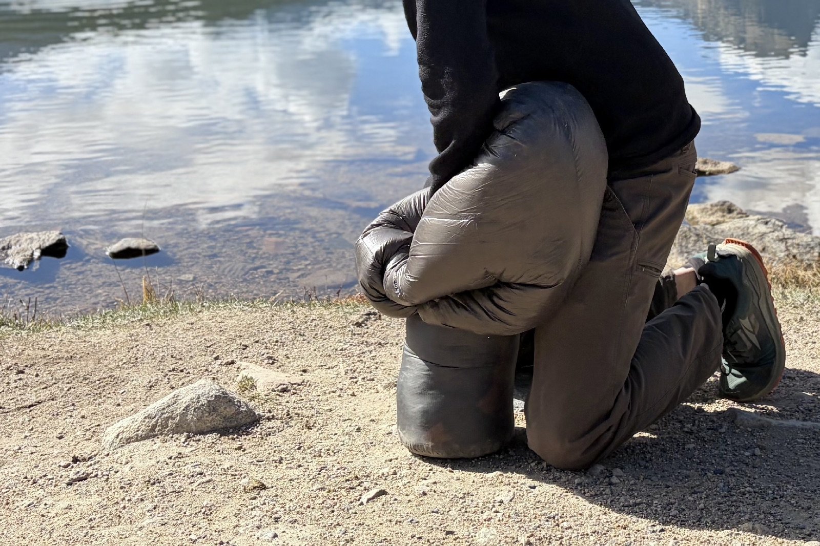 a hiker stuffs a grey sleeping quilt into a grey stuff sack by a lake