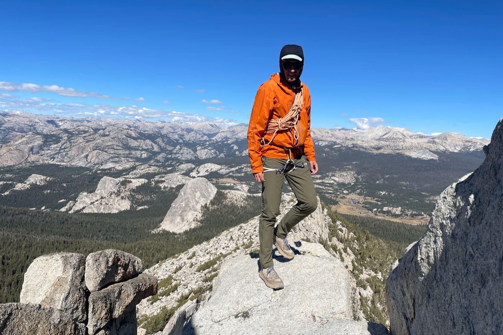 A man stands on a rocky summit with alpine views bhind him.