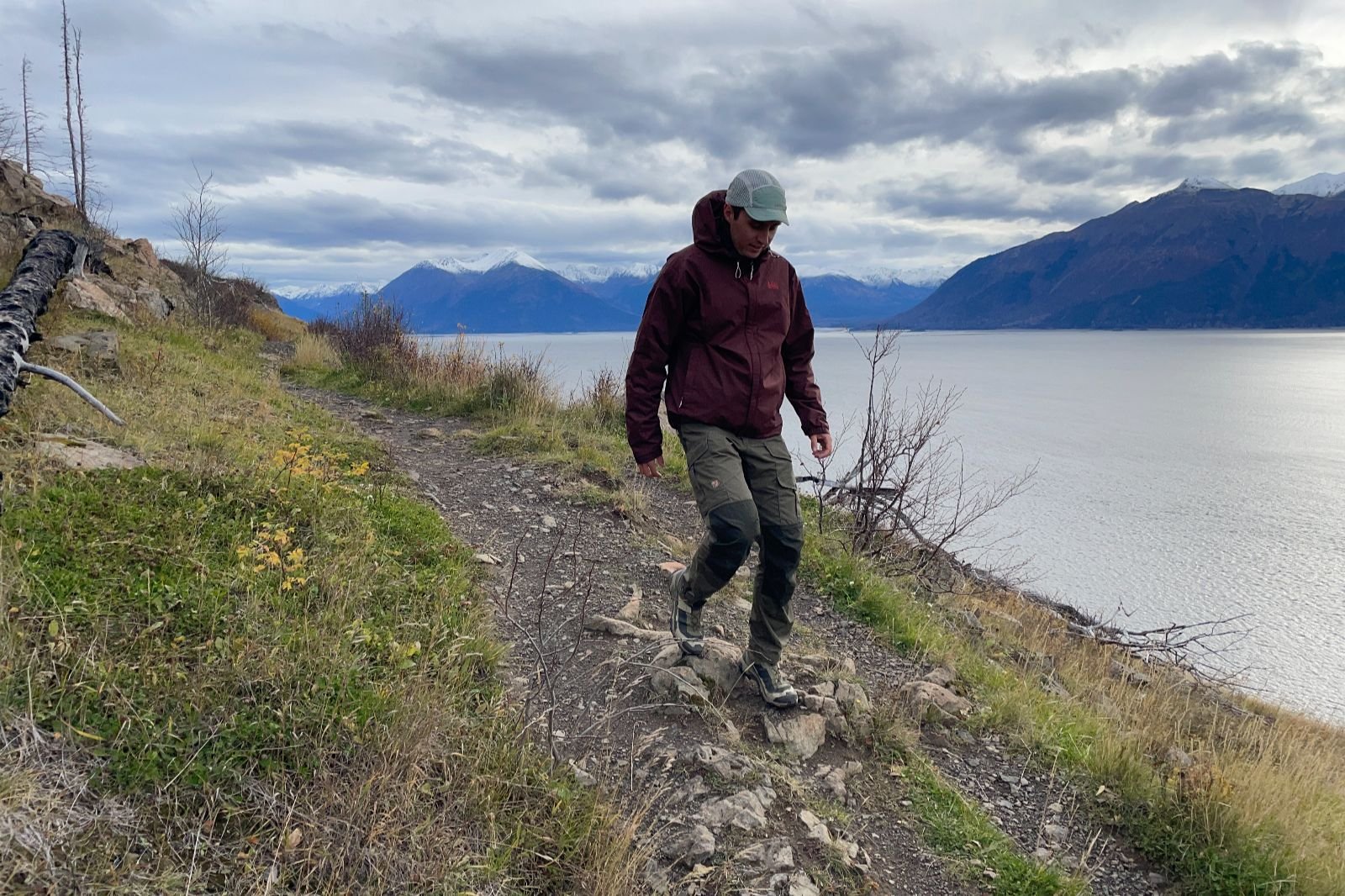 A man walks down a trail in front of an ocean and mountain view.