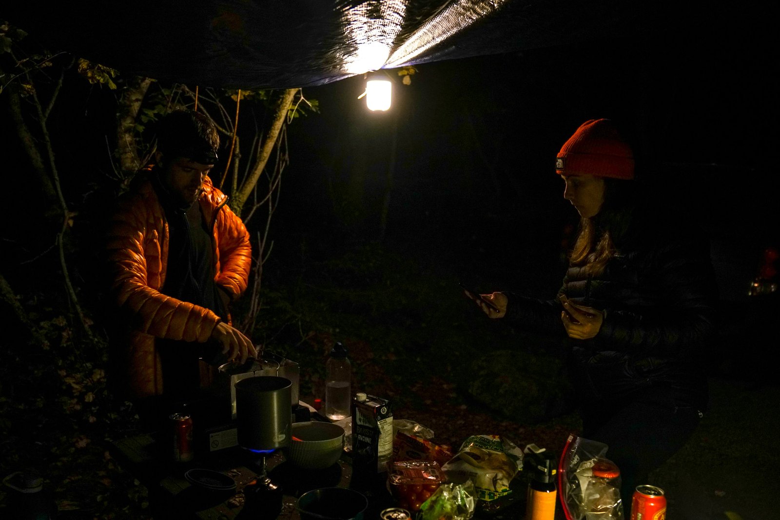 Two people stand around a picnic table covered in cooking gear in the dark. A tarp hangs above them with the BioLite AlpenGlow 500 lantern attached to its corner.