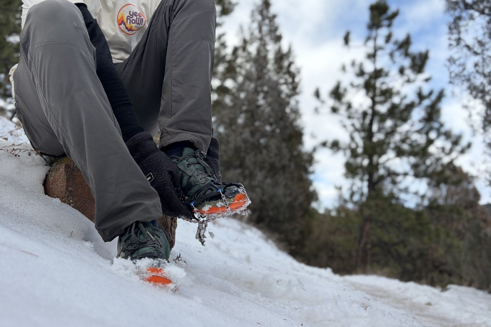 a hiker sits on a rock in the snow and puts on an ice cleat