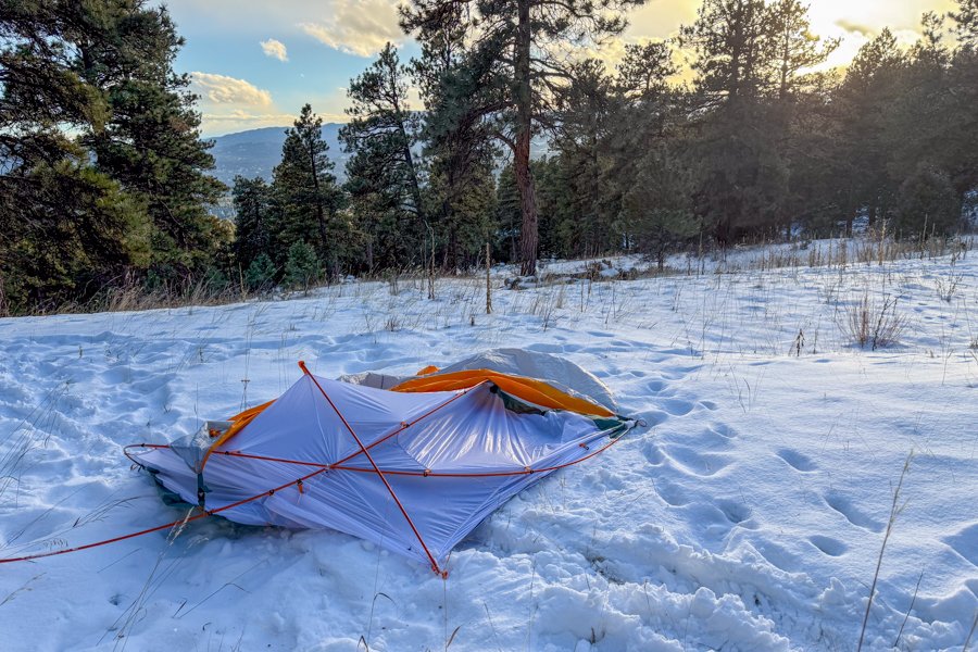 The Mineral King 2 All Season tent collapsed flat under snow load, with poles splayed outward on a snowy hillside surrounded by pine trees at sunset.